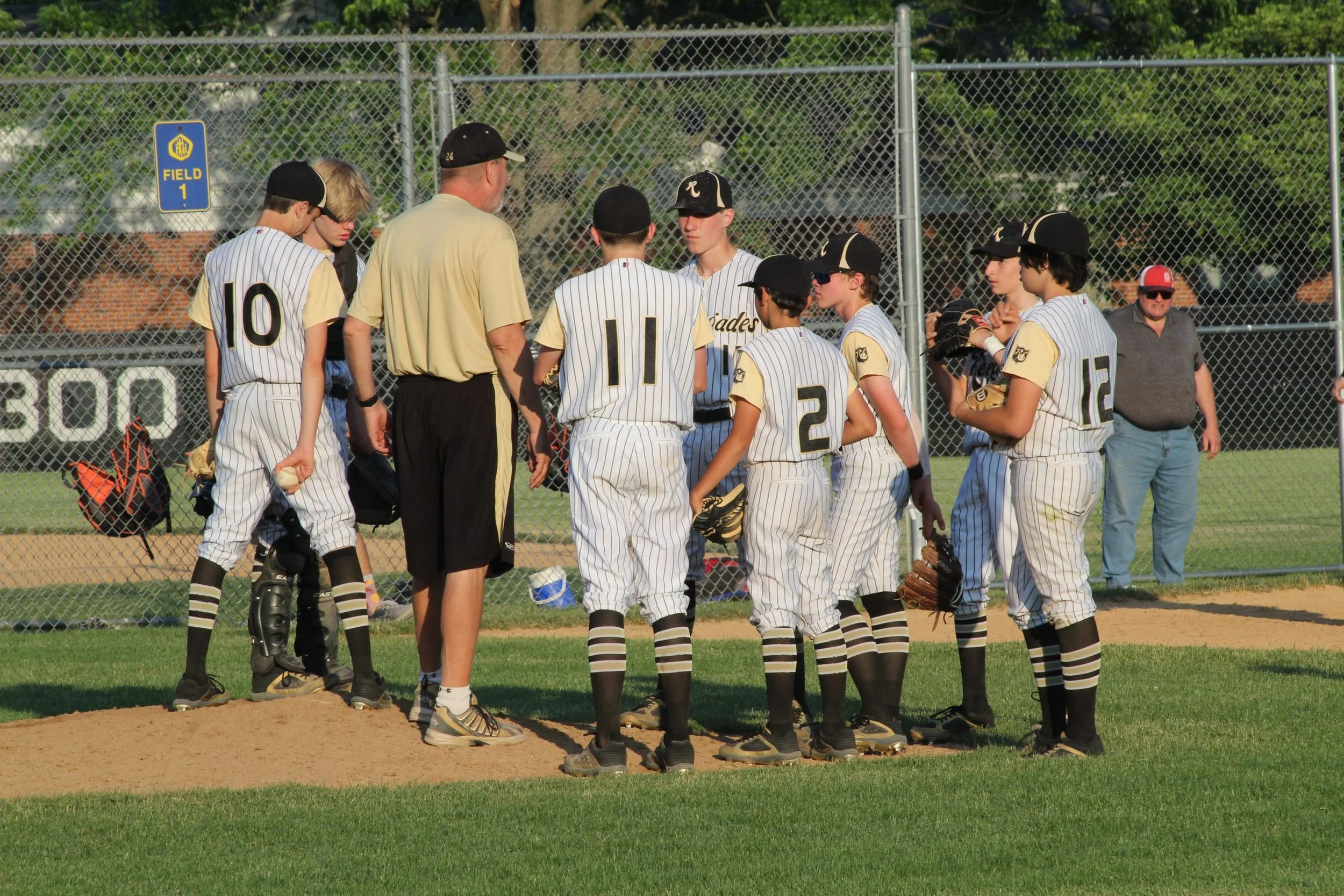 Youth baseball team in a huddle on the field during a game, with coaches and spectators in the background.