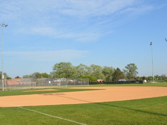 Empty baseball field with green grass, brown dirt infield, and a chain-link fence, under a blue sky with a few clouds.
