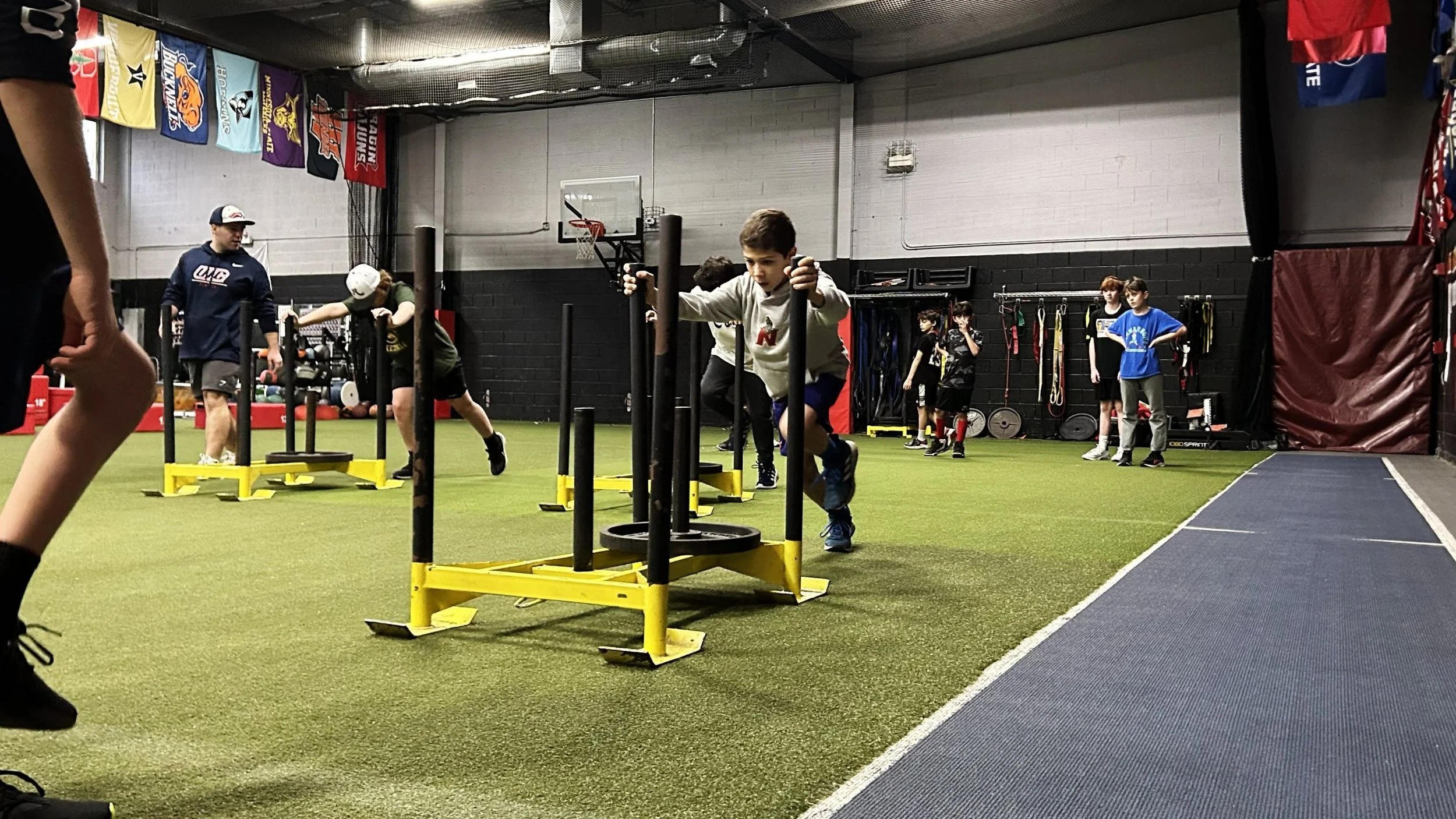 Young boys participating in a strength and agility training session at an indoor gym, with some pushing sleds and others waiting in line.