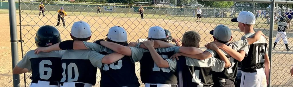 Youth baseball team members in black and gray uniforms and helmets watching a game from behind a chain-link fence, with players on the field in the background.