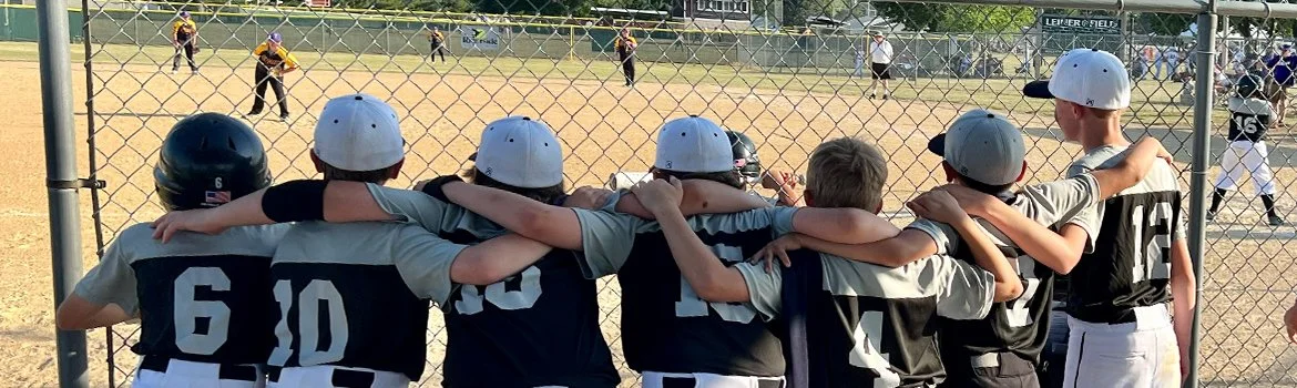 Young baseball players wearing helmets and team uniforms, with arms around each other, watching a game on the field.