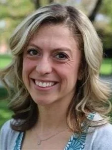 A smiling woman outdoors with trees and sunlight in the background.