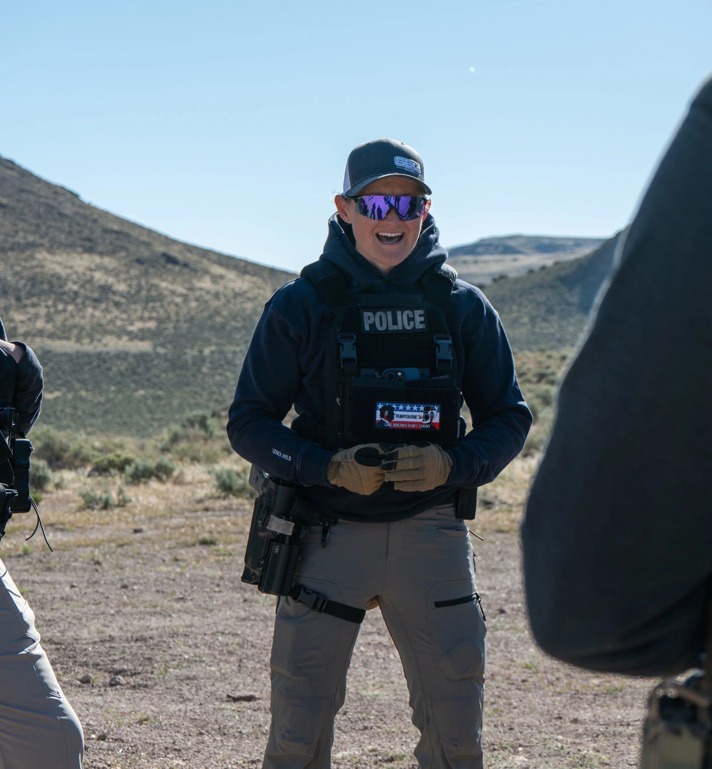 A police officer wearing tactical gear, including a bulletproof vest labeled 'POLICE,' standing outdoors in a desert landscape with mountains in the background. The officer has sunglasses, a cap, and is smiling while holding a device.