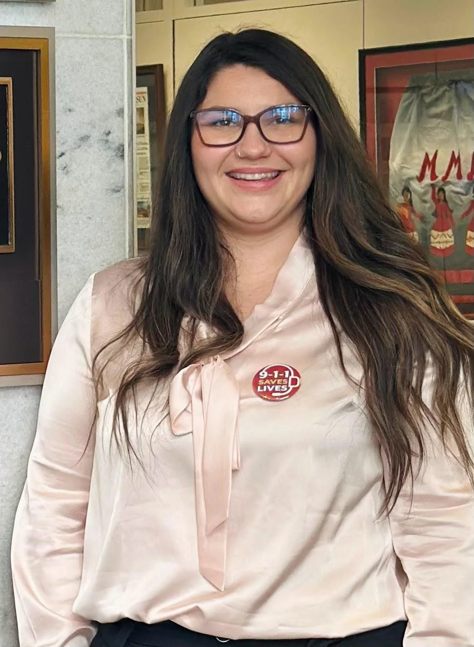 A woman with long dark hair, glasses, and a nose piercings smiling at the camera. She is wearing a light pink satin blouse with a tie neck and a button that reads '3-1-1 Saves Lives.' She is indoors with framed pictures and posters on the wall behind her.