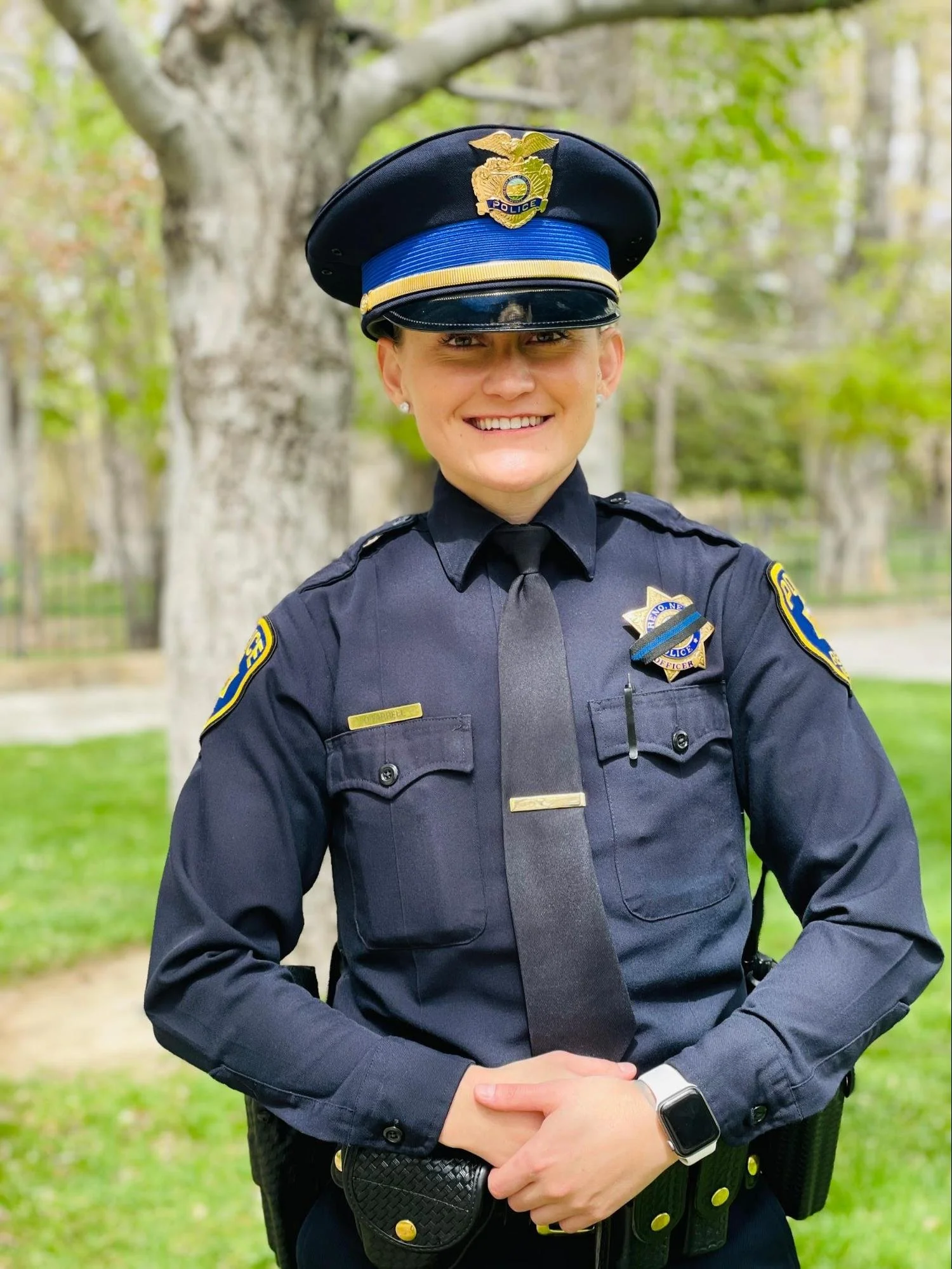 A smiling female police officer in uniform standing outdoors with green trees in the background.