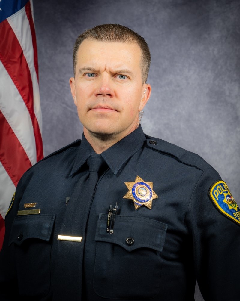 Portrait of a police officer in uniform, standing next to an American flag, with a serious expression and a gray background.