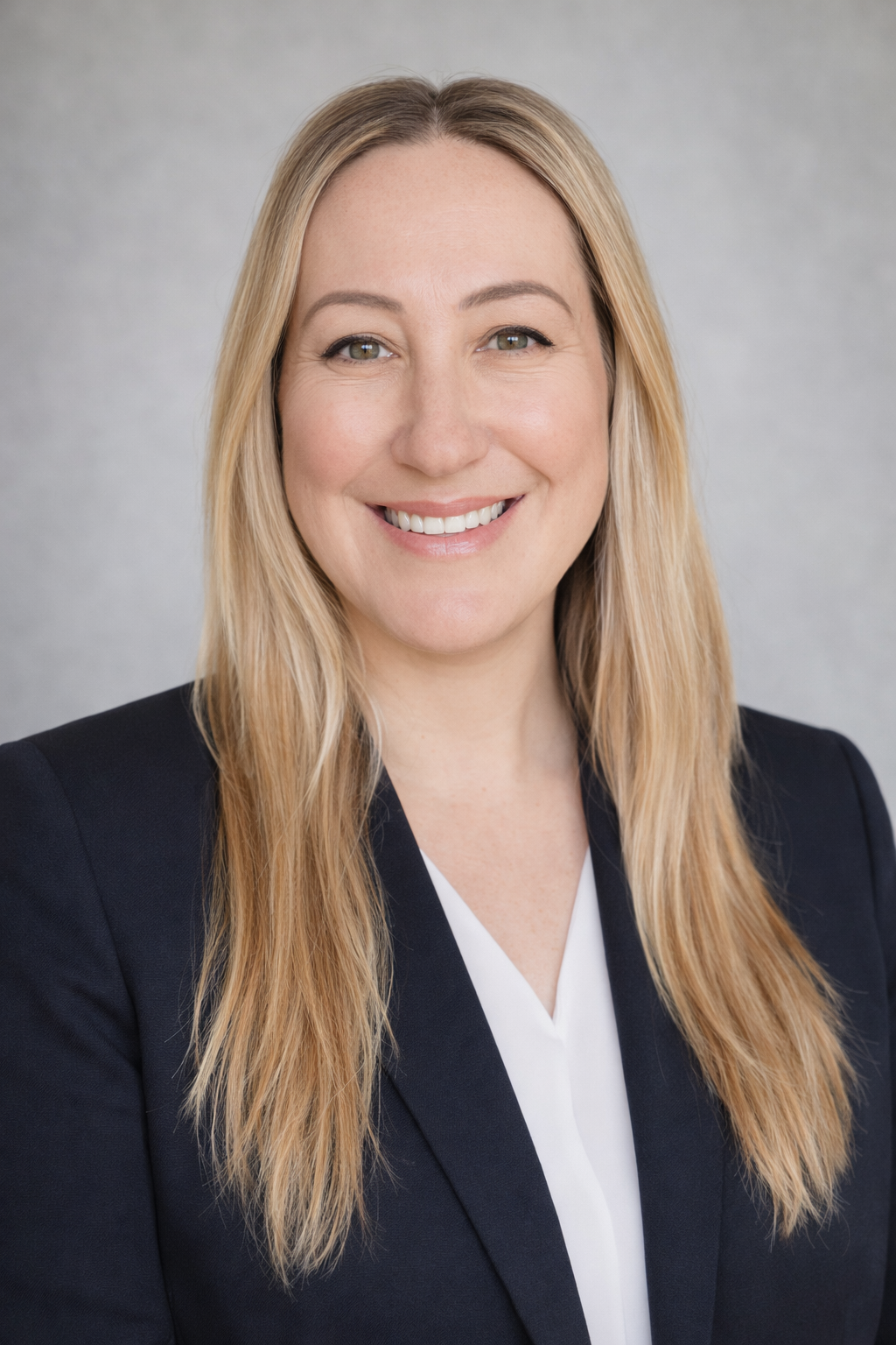 Professional headshot of a smiling woman with long blonde hair, wearing a dark blazer and white blouse, against a plain gray background.