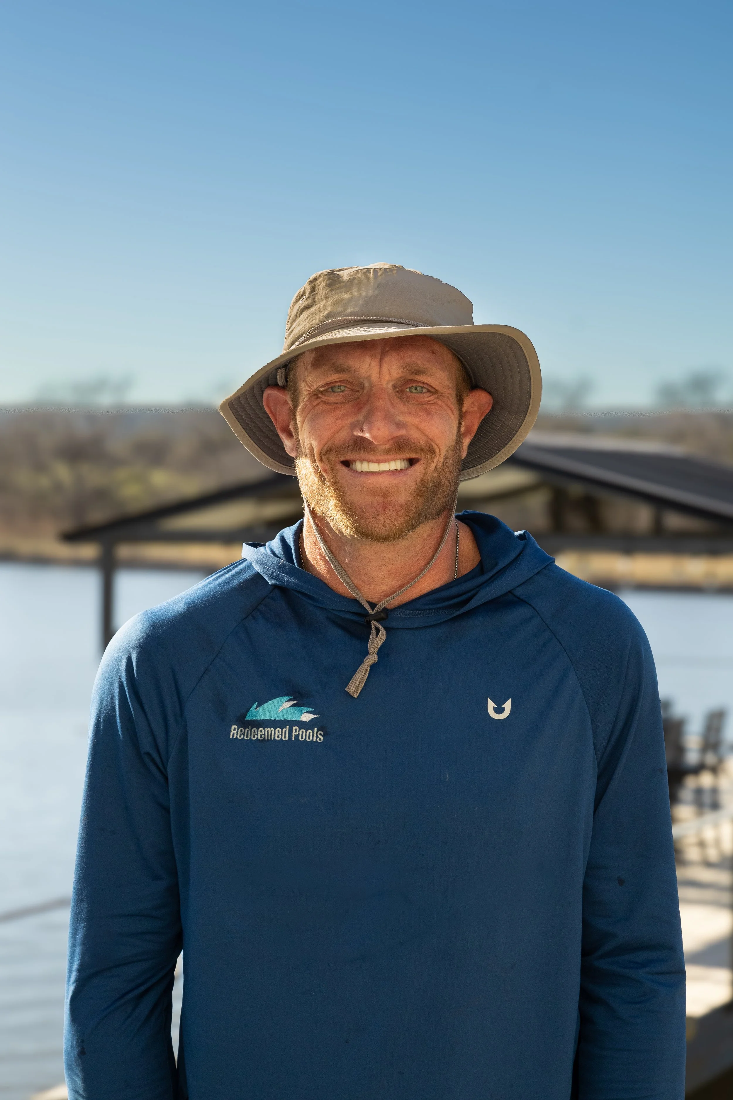 Smiling man outdoors near water, wearing a wide-brimmed hat and a blue hoodie with 'Redeemed Pools' logo, background shows a shoreline and a covered wooden dock.