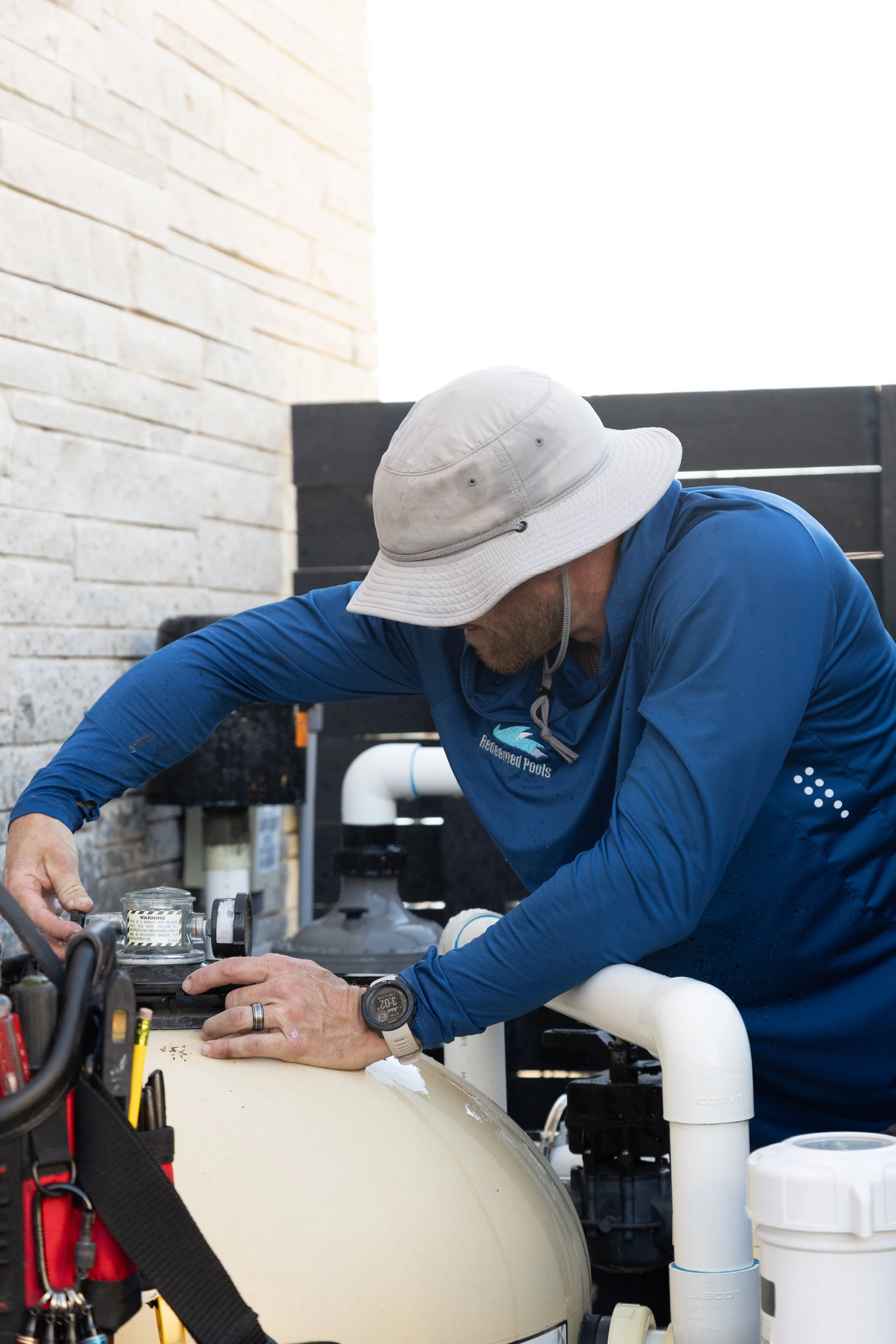 A man wearing a blue long-sleeve shirt and a white bucket hat working on pool equipment outdoors.