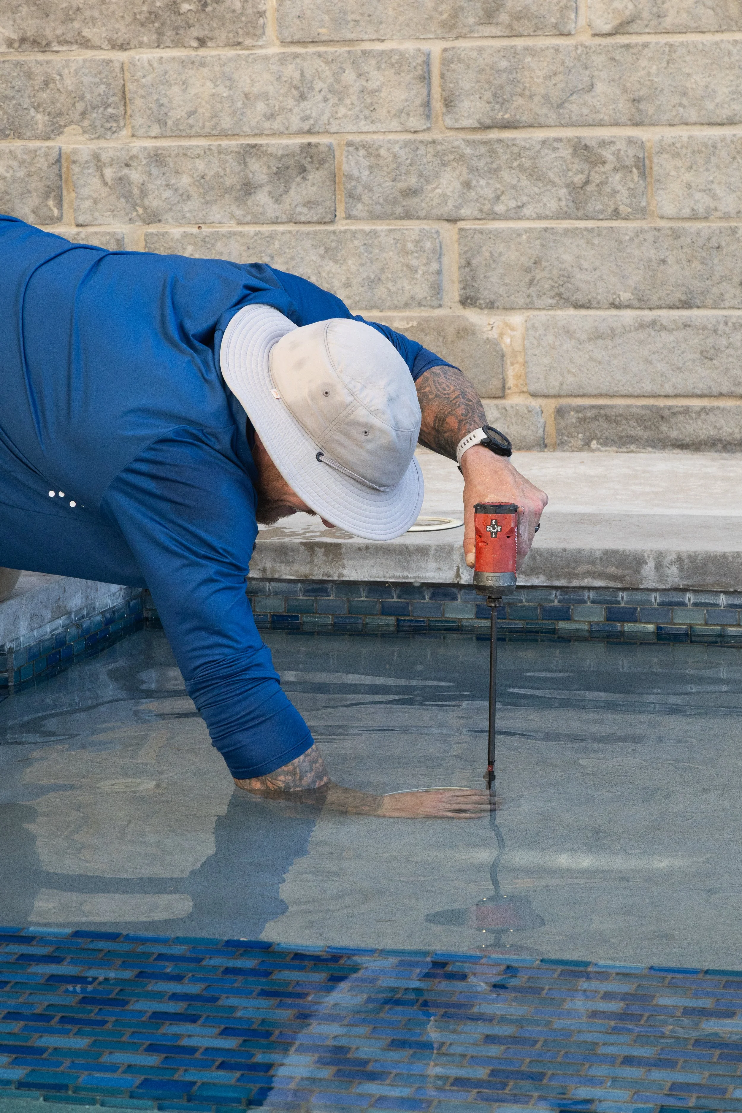 A man wearing a large white hat and a blue long-sleeve shirt is leaning over the edge of a swimming pool, using a tool to work on the pool's surface.