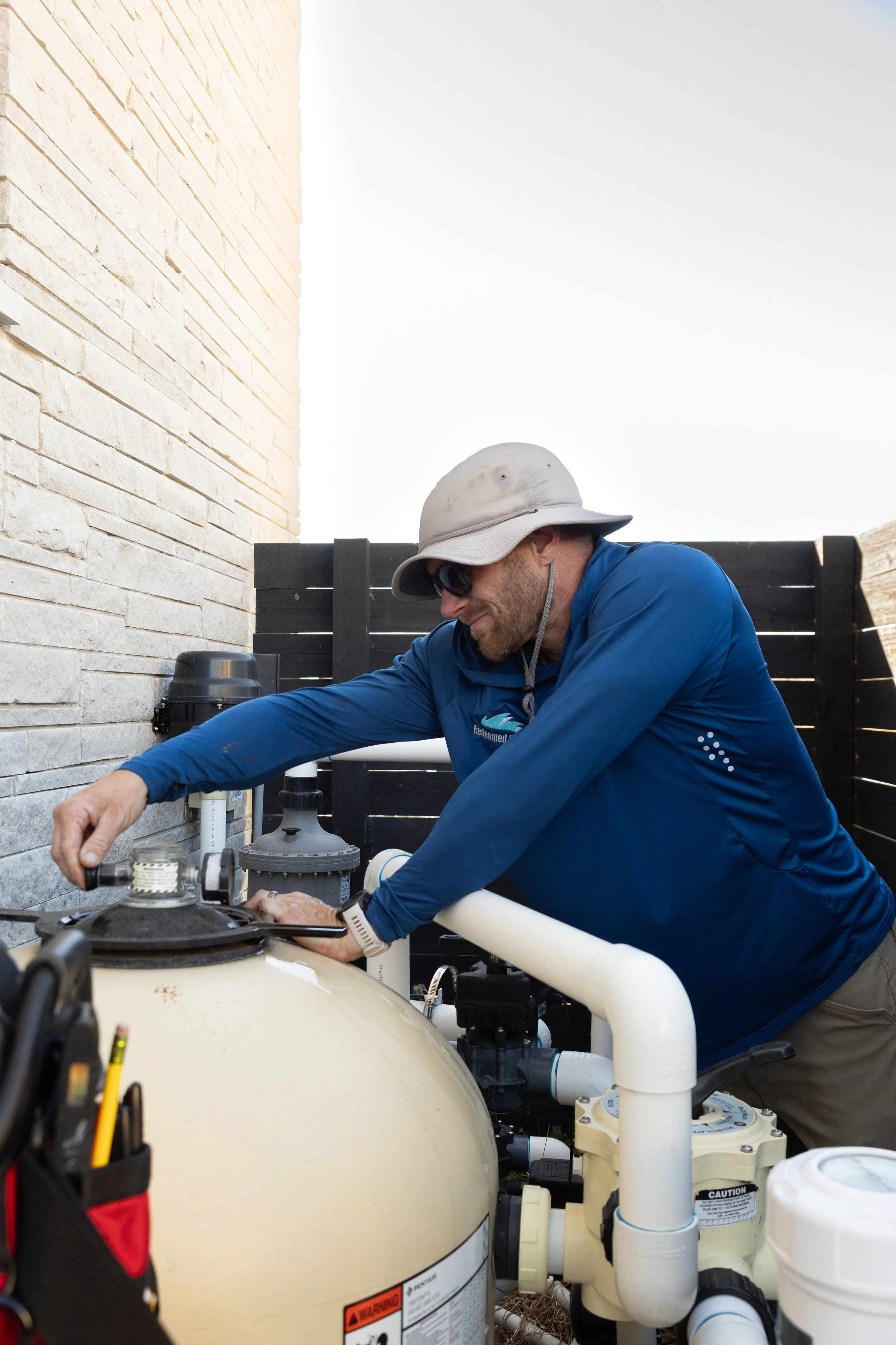 A man wearing a bucket hat, sunglasses, and a blue long-sleeve shirt is adjusting equipment outdoors near a pool or water system.