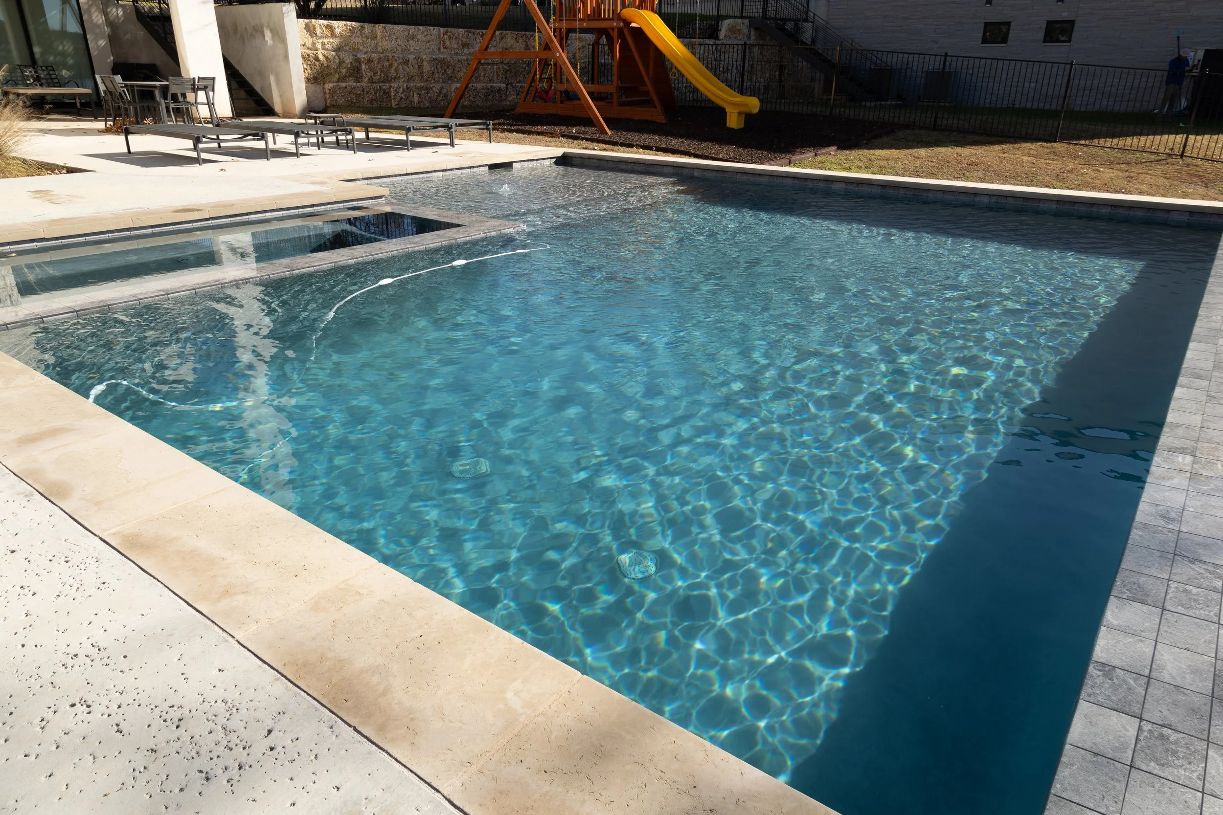 A modern backyard swimming pool with a hot tub and a yellow slide, surrounded by concrete and dark paving stones, with outdoor furniture and a playground in the background.