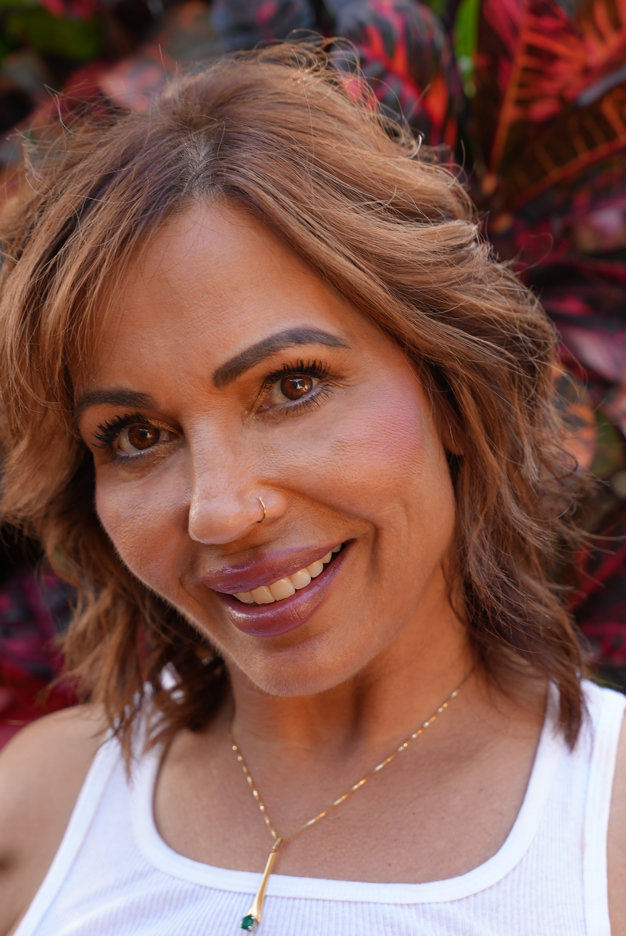 Close-up of a smiling woman with brown hair, brown eyes, a nose piercing, wearing a white tank top and a gold necklace with a green pendant, standing outdoors against a background of purple and green foliage.