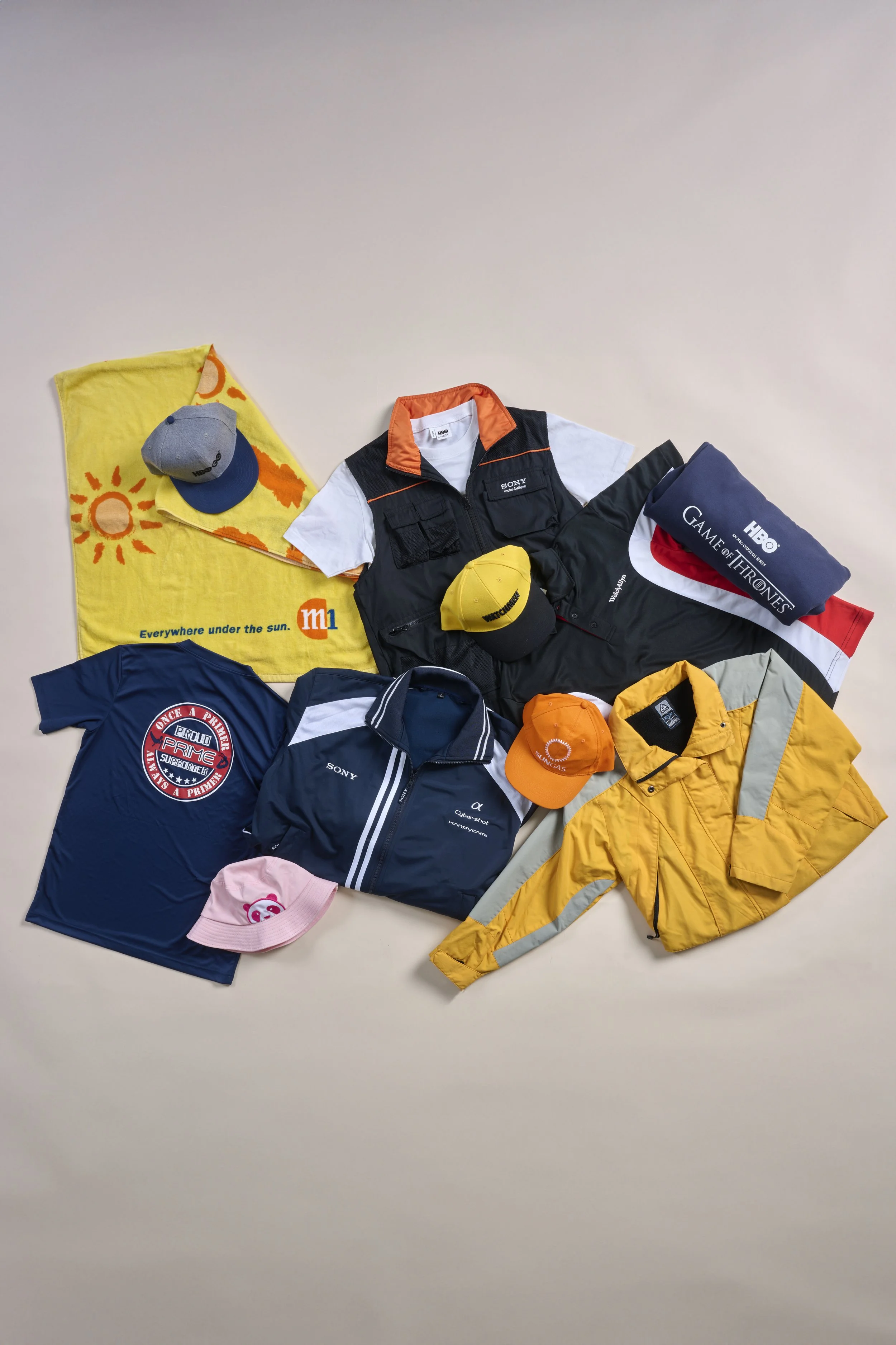 Various clothing items and accessories laid out on a white surface, including jackets, caps, t-shirts, and a towel, with some logos and graphic designs visible.