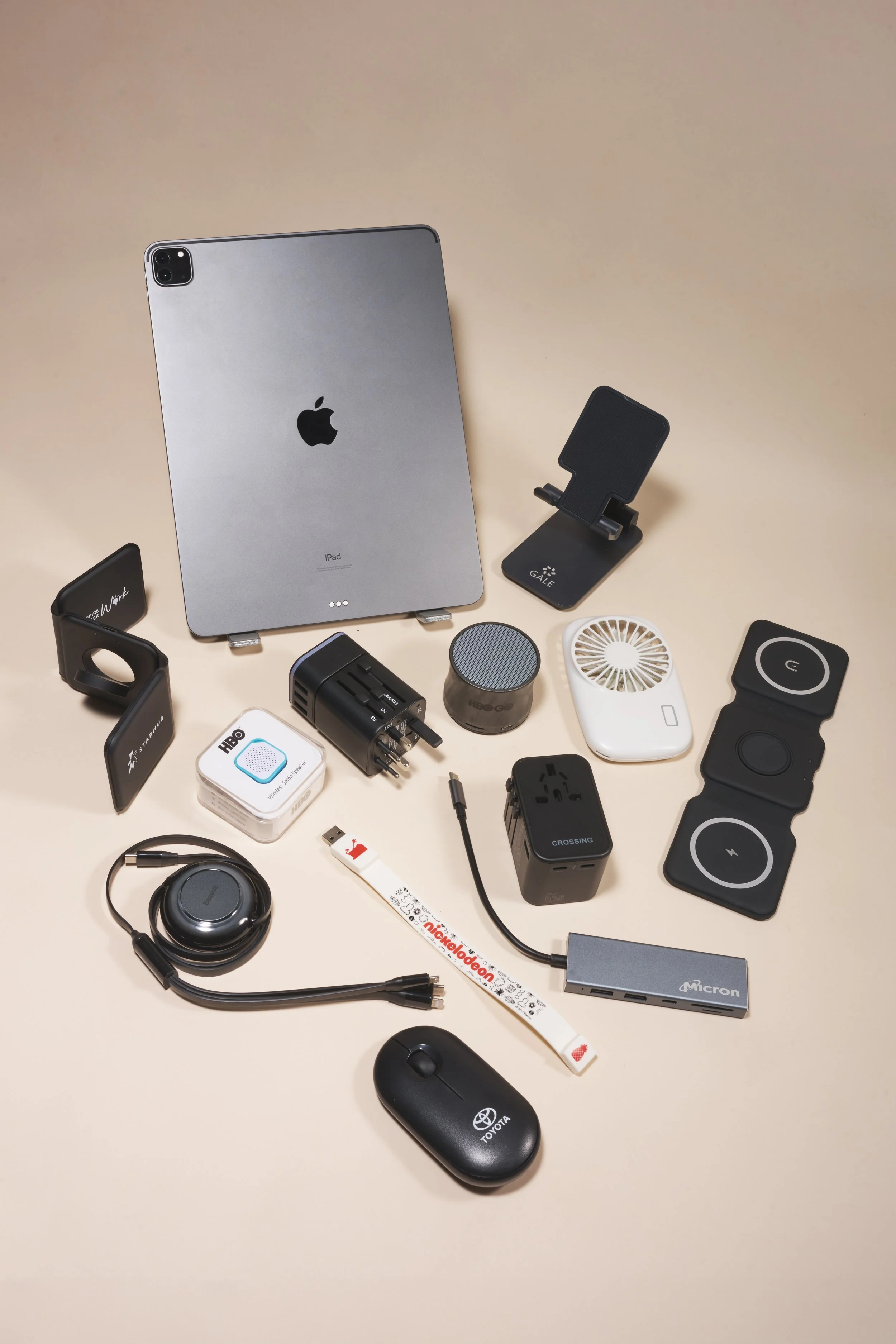 Flat lay of various electronic devices and accessories including an iPad, wireless chargers, a mouse, a portable speaker, a power strip, a headset, a USB flash drive, an adapter, a compact fan, and other small electronic gadgets on a beige background.