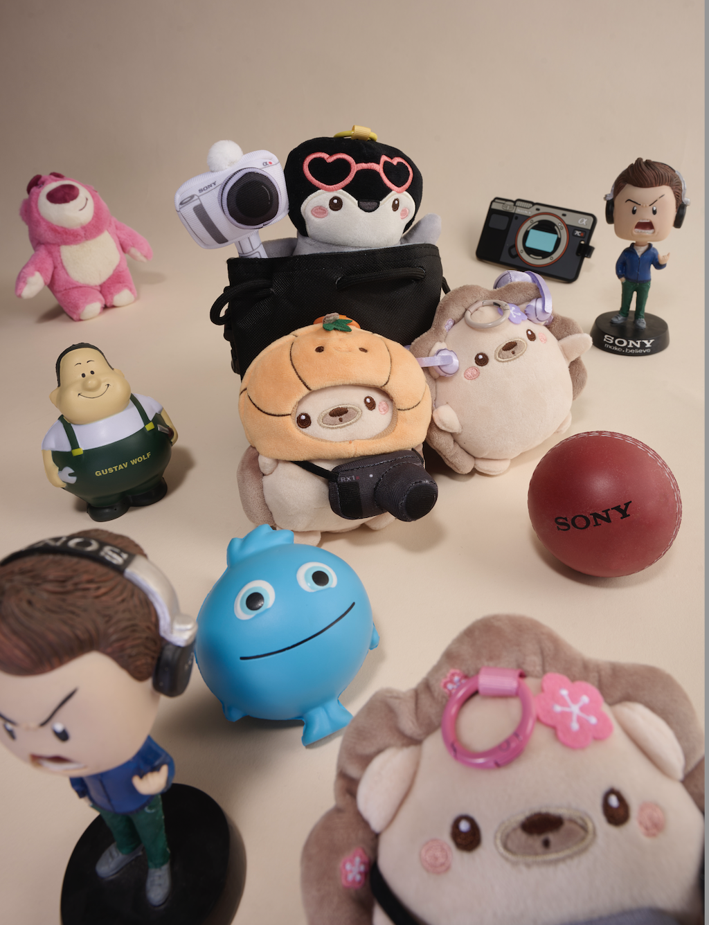Collection of plush toys, action figures, and miniature cameras on a beige surface, featuring characters with headphones, cameras, and a Sony ball, with a plain background.
