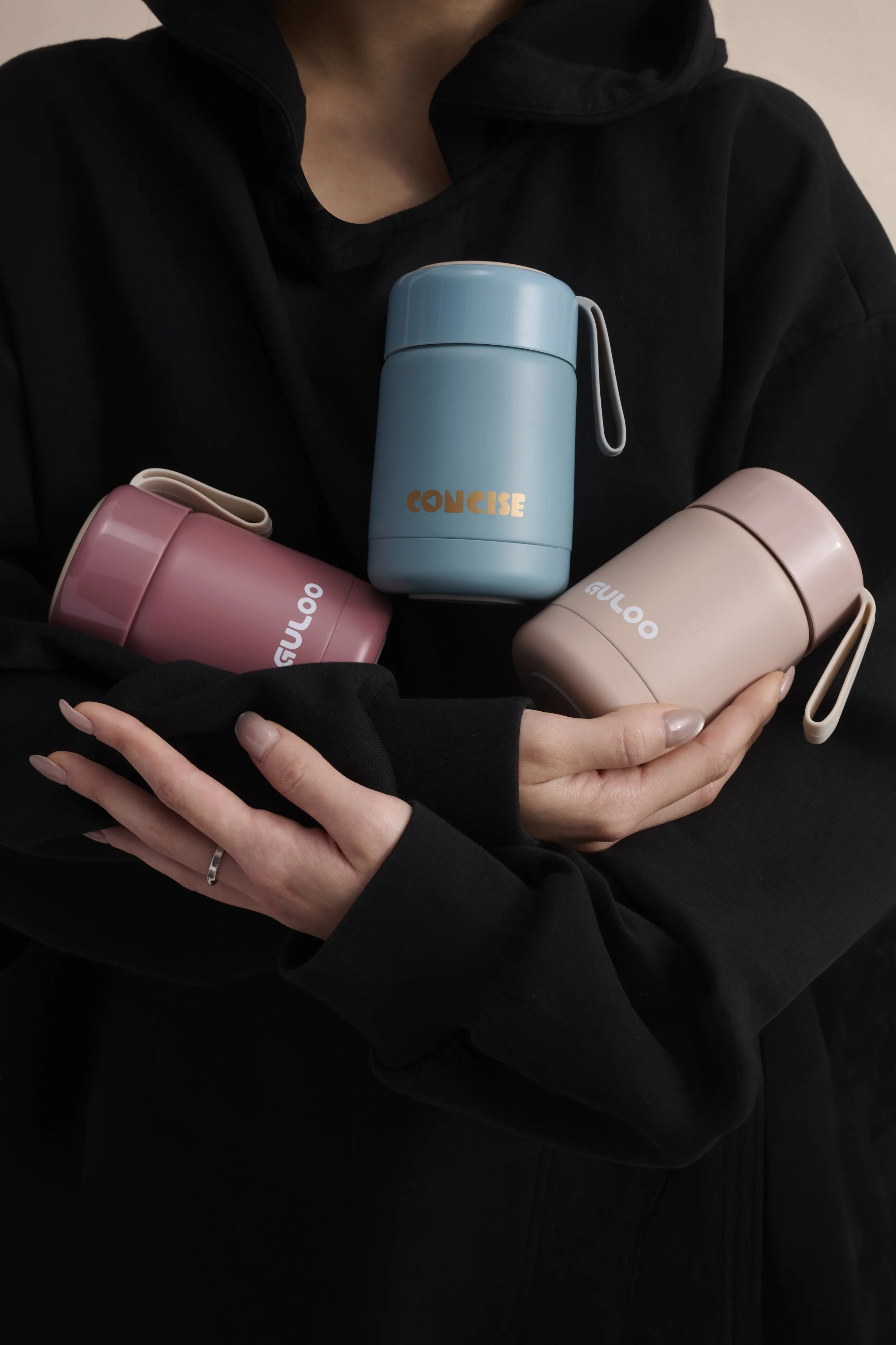 Person holding three pastel-colored insulated travel mugs, one blue with 'CONCISE' text, and two pinks with 'GULOO' text, against a black hoodie background.