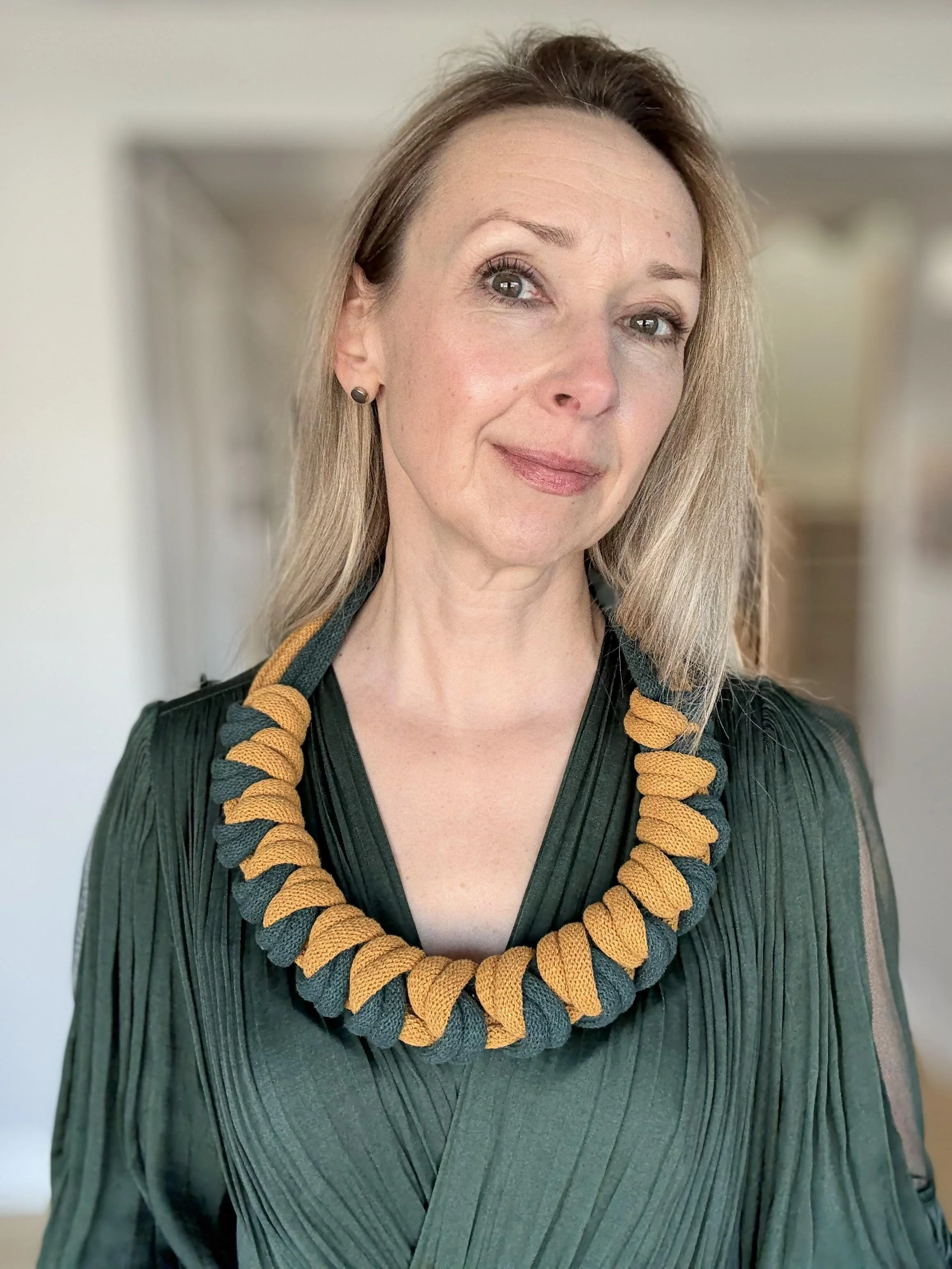 A woman with light brown hair, fair skin, and gray eyes, wearing a dark green pleated dress and a chunky necklace made of dark green and yellow braided rope.