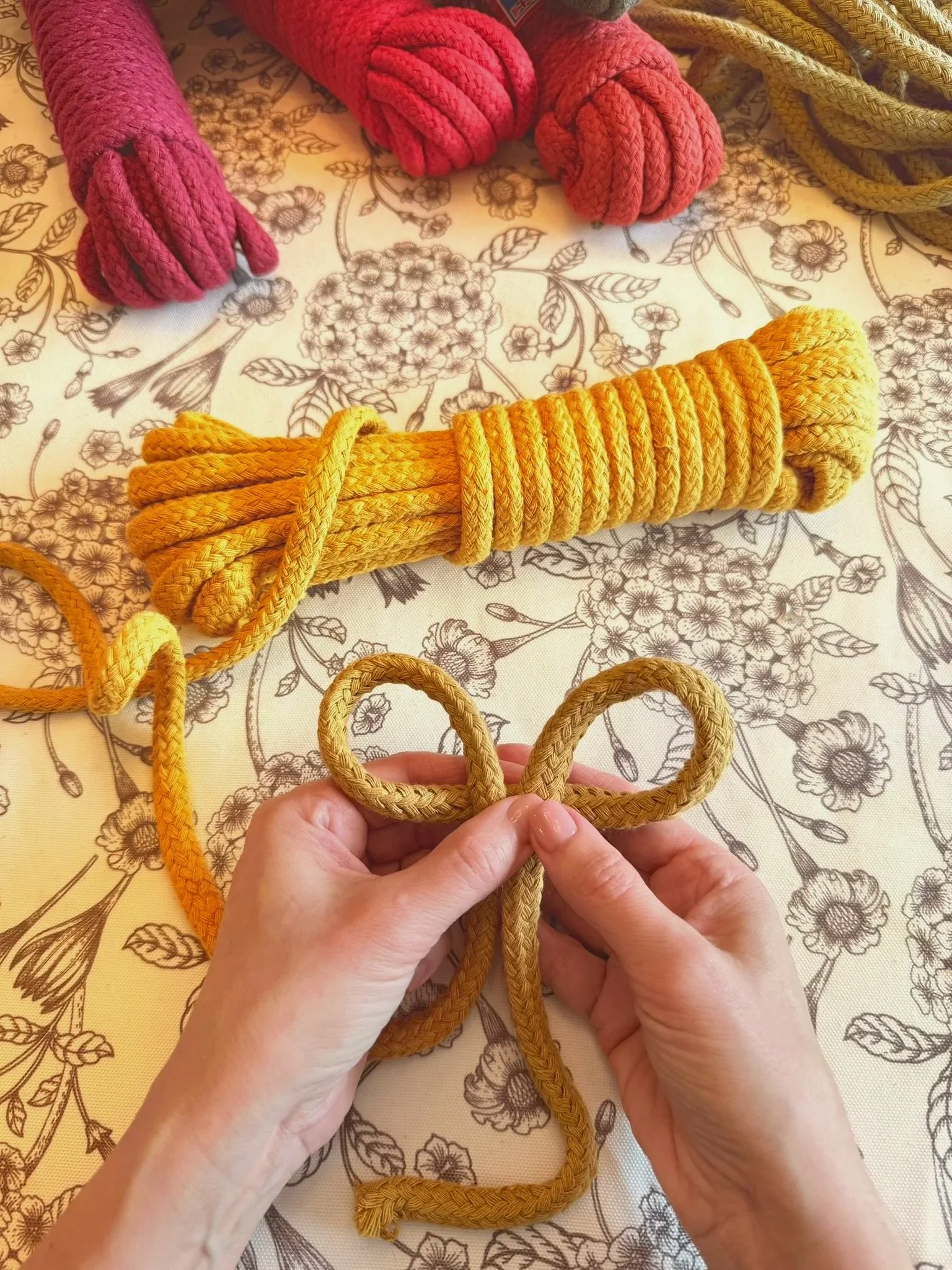 Person tying a yellow rope into a decorative bow on a floral tablecloth with additional ropes in pink, red, and green in the background.