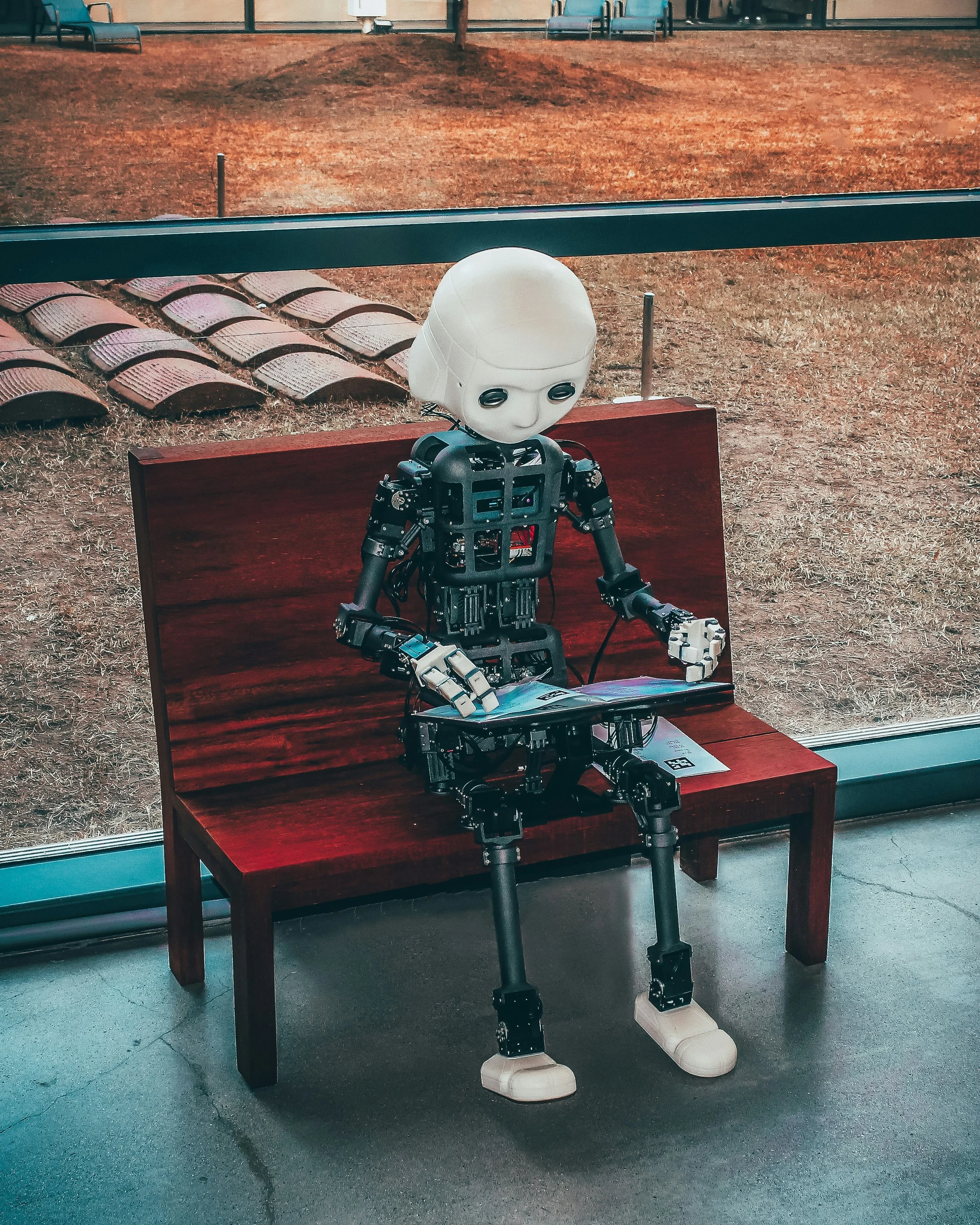 Robot with a humanoid head sitting on a wooden bench indoors, looking at a tablet, with a glass window showing grass and a bike rack outside.