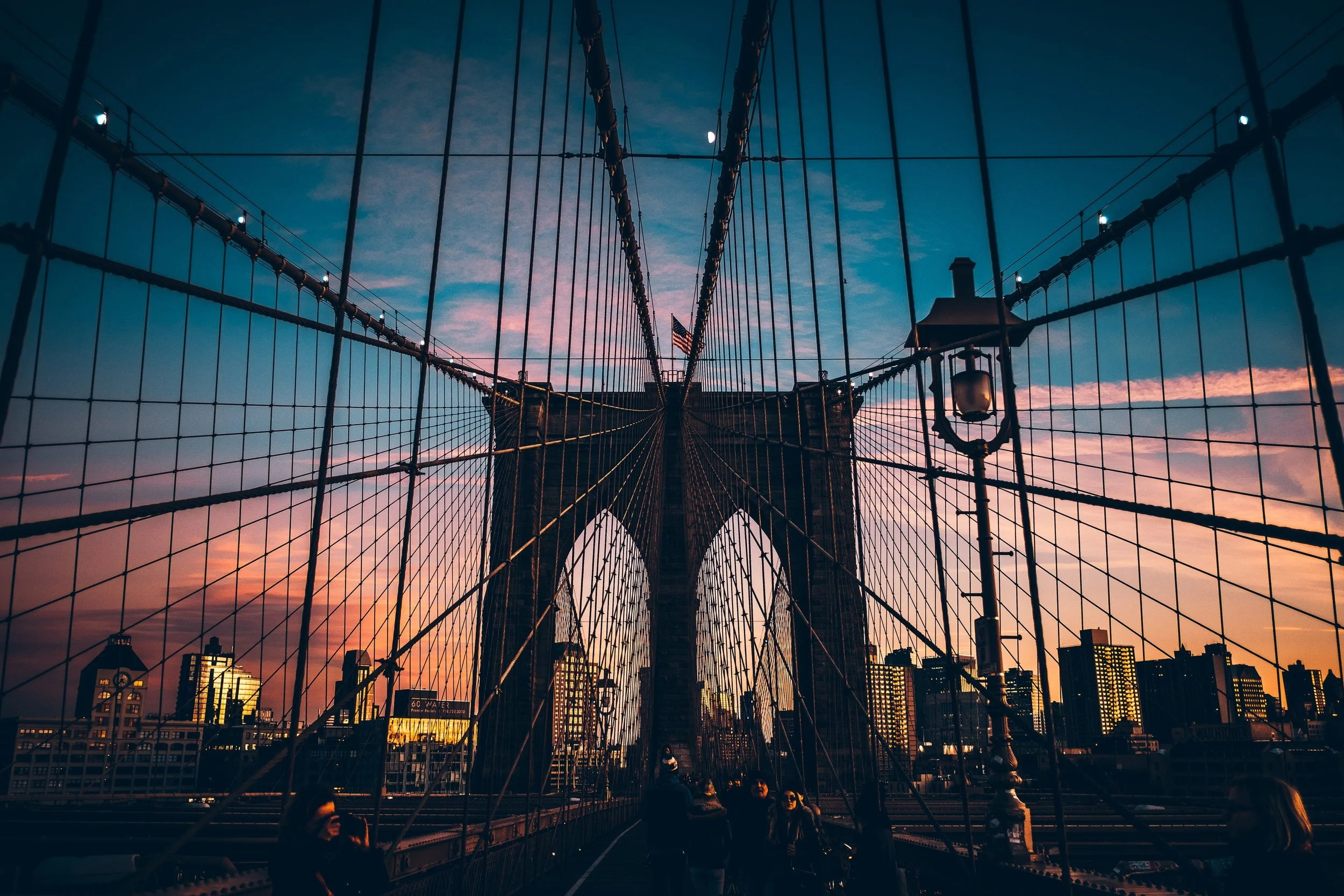 View of Brooklyn Bridge at sunset with silhouetted pedestrians, city skyline, and sky with orange and pink hues.