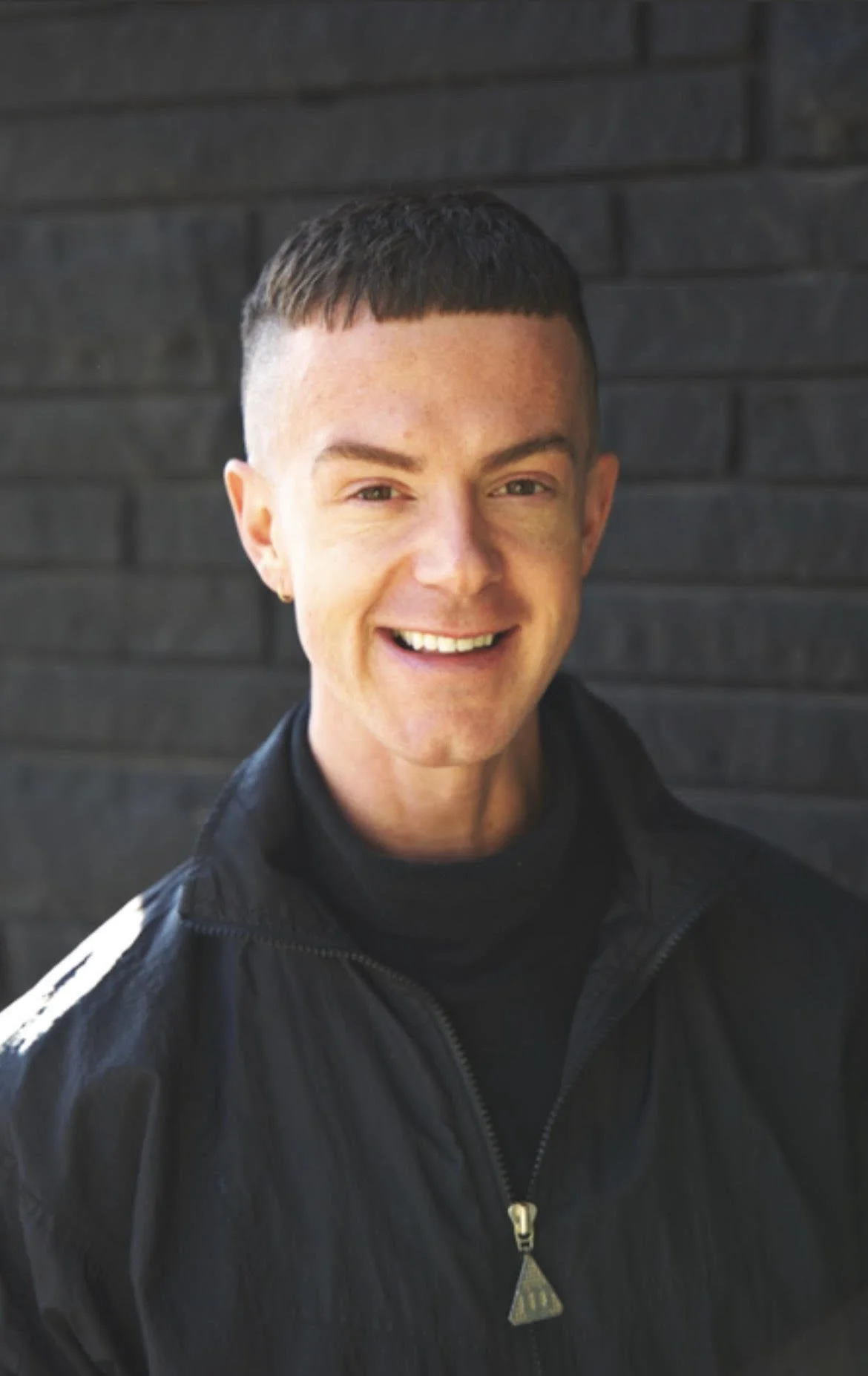 McKay Gordon smiles in headshot wearing black jacket against a black wall. White non-binary person in their 30's smiling. Brown hair, hazel eyes.
