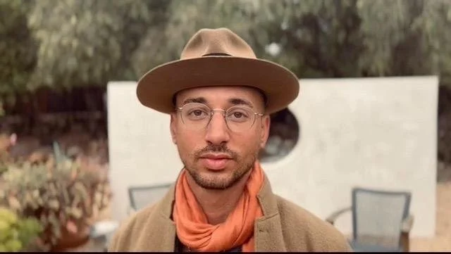 A man wearing glasses, a tan wide-brimmed hat, an orange scarf, and a brown coat portrait outdoors with a white brick structure and chairs in the background.