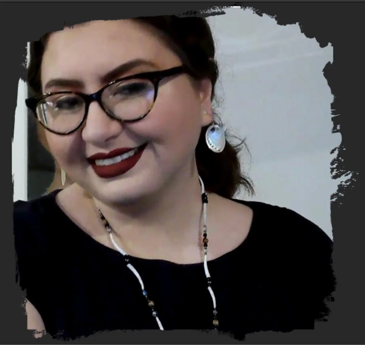 A woman with glasses, red lipstick, earrings, necklace, smiling and looking at the camera.