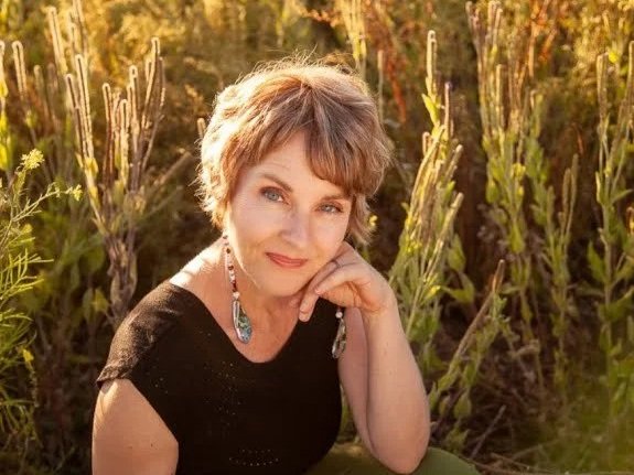 A woman with short gray hair sitting outdoors among tall plants during sunset, wearing a black top and a necklace, smiling softly.