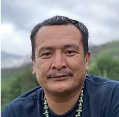 A close-up of a man outdoors with greenery and cloudy sky in the background.