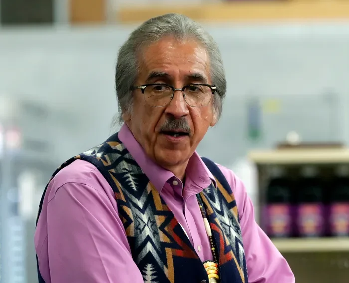Older man with gray hair and glasses, wearing a pink shirt and a colorful patterned vest, standing in a store or warehouse.