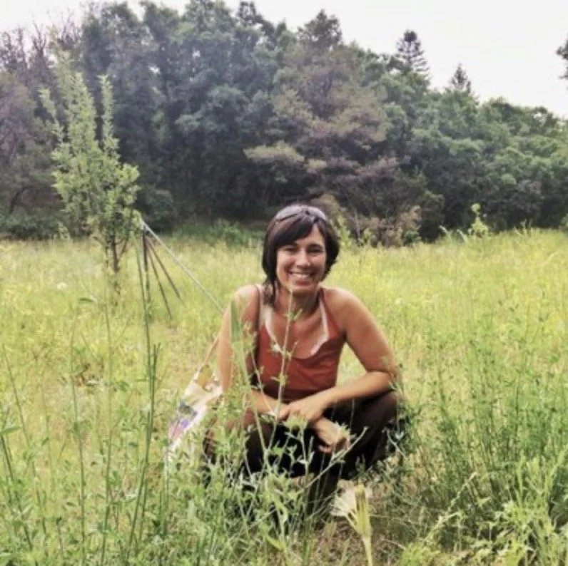 A woman crouching in a grassy field with trees in the background, smiling at the camera.