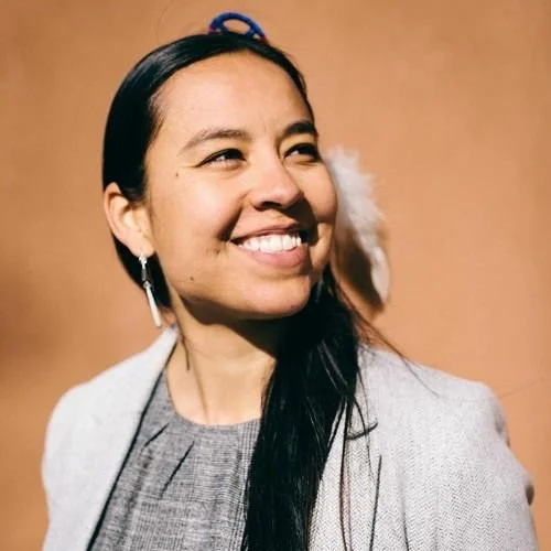 A smiling woman with long dark hair, wearing a gray blazer and earrings, standing against a plain brown background.