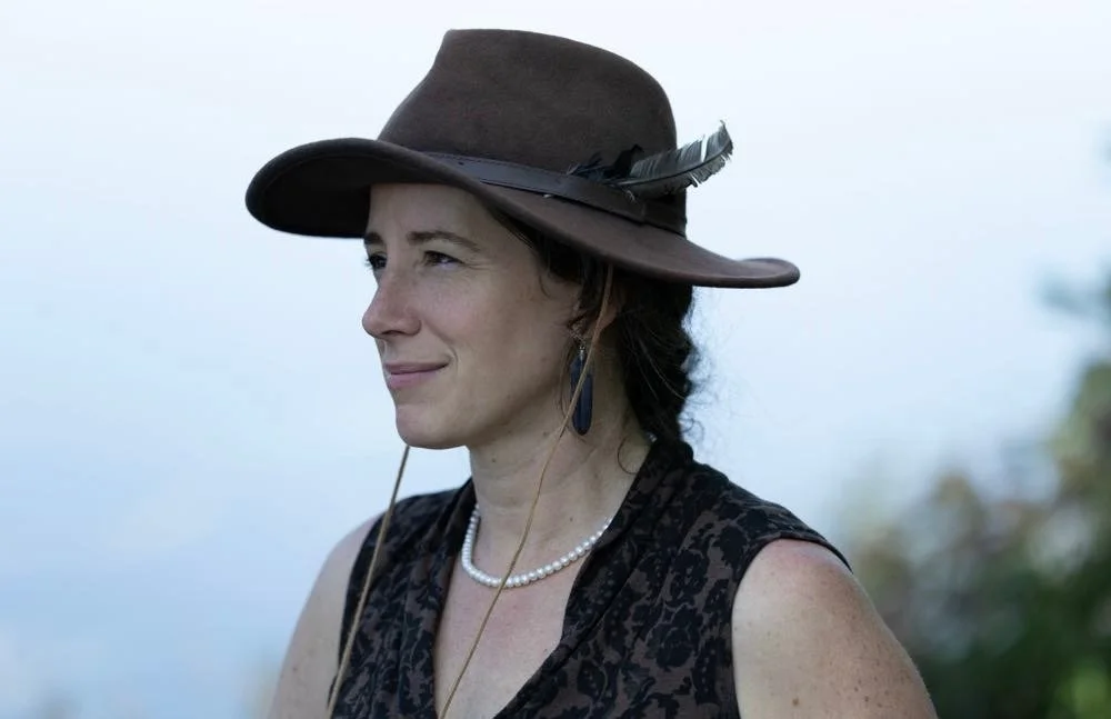 Woman wearing a hat with a feather in its band in front of a misty lake 