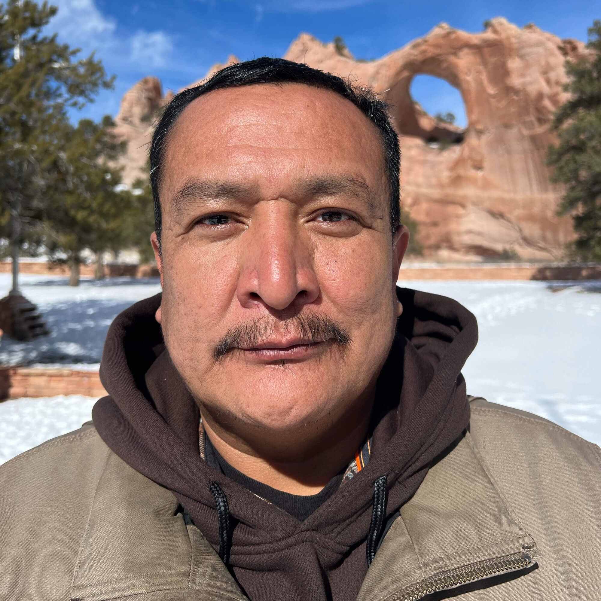 A man with short dark hair and a necklace outdoors with a cloudy sky and trees in the background.
