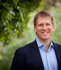 A smiling man with light brown hair wearing a dark blazer and light blue shirt outdoors with green trees in the background.