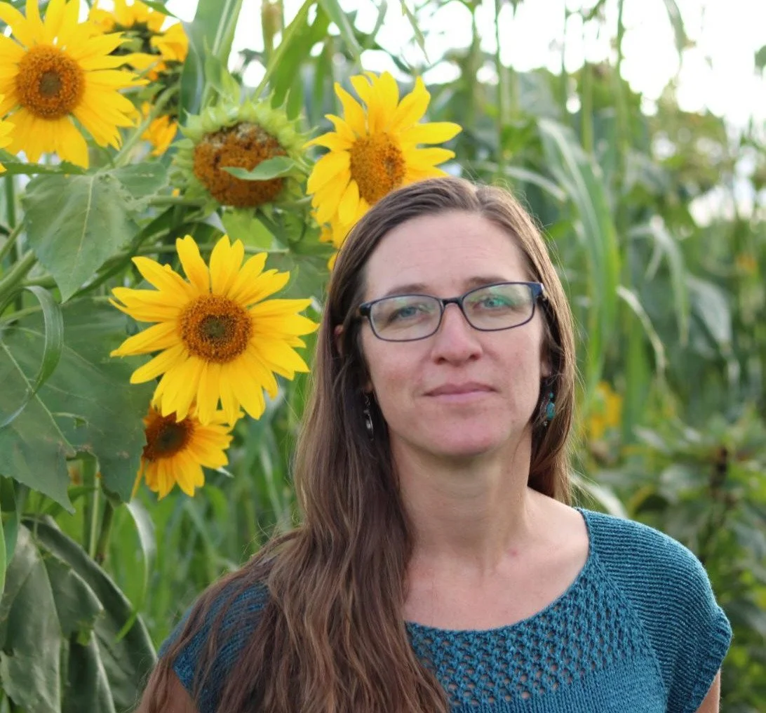 Woman with long brown hair and wire-rimmed glasses standing in a sunflower field