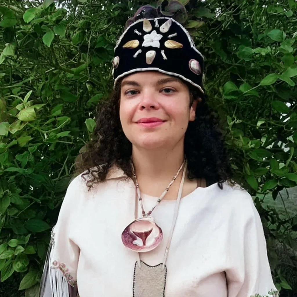 Woman wearing a black hat decorated with shells and a large necklace with a pink shell pendant, standing in front of green shrubbery.