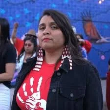 A woman with dark hair wearing a red shirt with a white handprint design and a black jacket. She is outdoors, with several people and a colorful mural in the background.