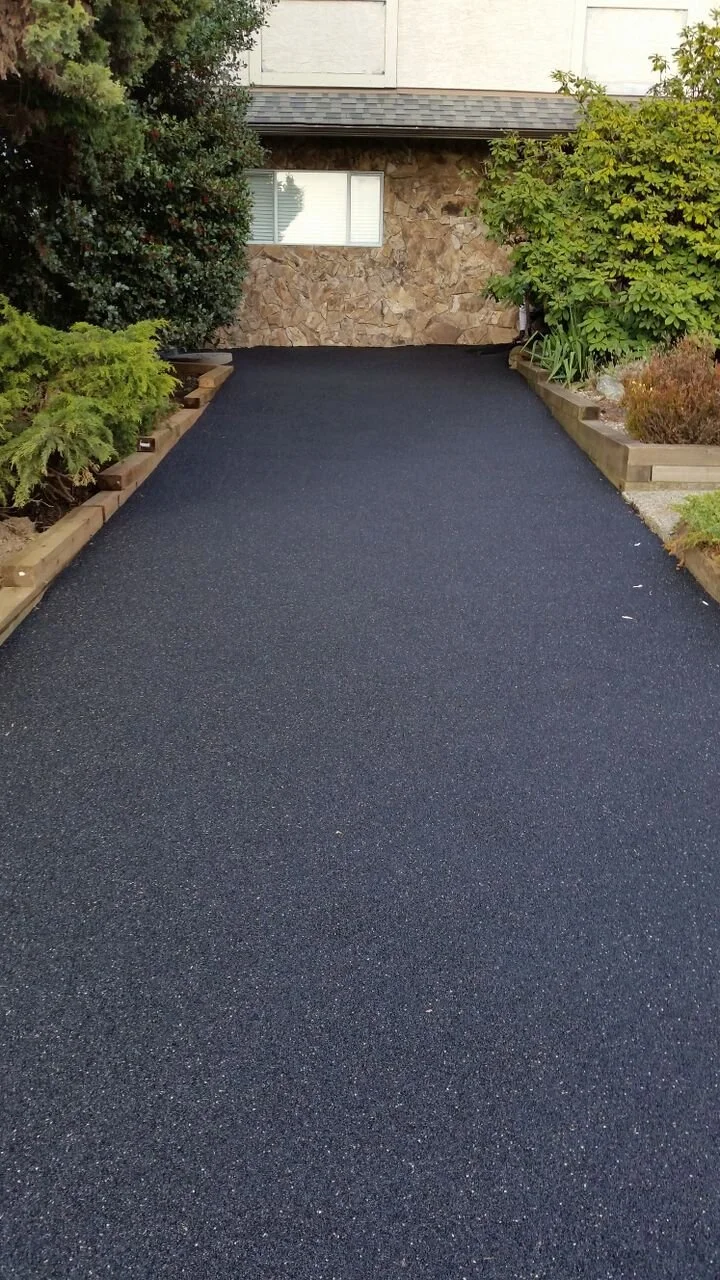 Freshly paved black asphalt driveway leading to a house with a stone facade, bordered by green bushes and plants.