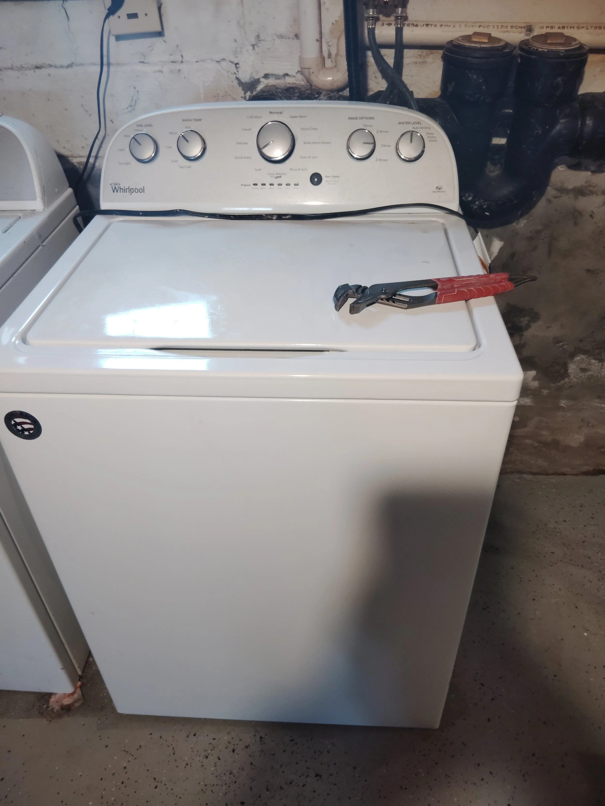 A white top-loading washing machine with a pair of pliers resting on the lid, situated in a basement setting with exposed brick wall and pipes overhead.