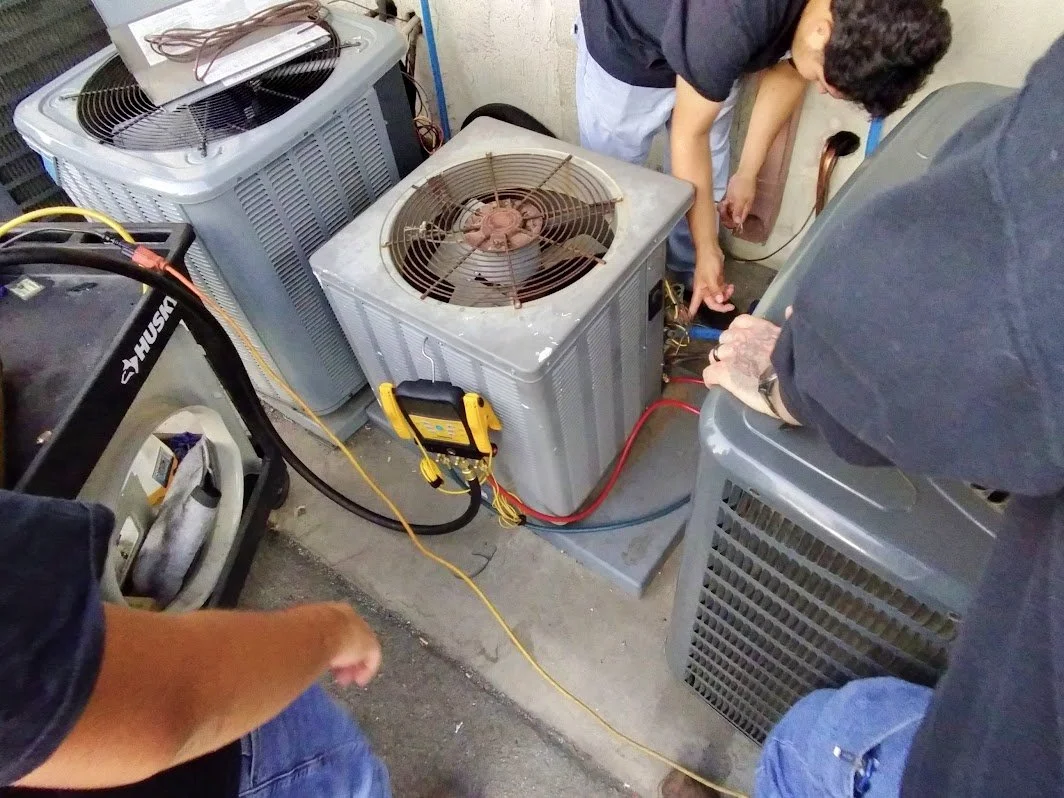HVACR technicians repairing or servicing an air conditioning unit with multiple outdoor compressor units, tools, and electrical wiring.