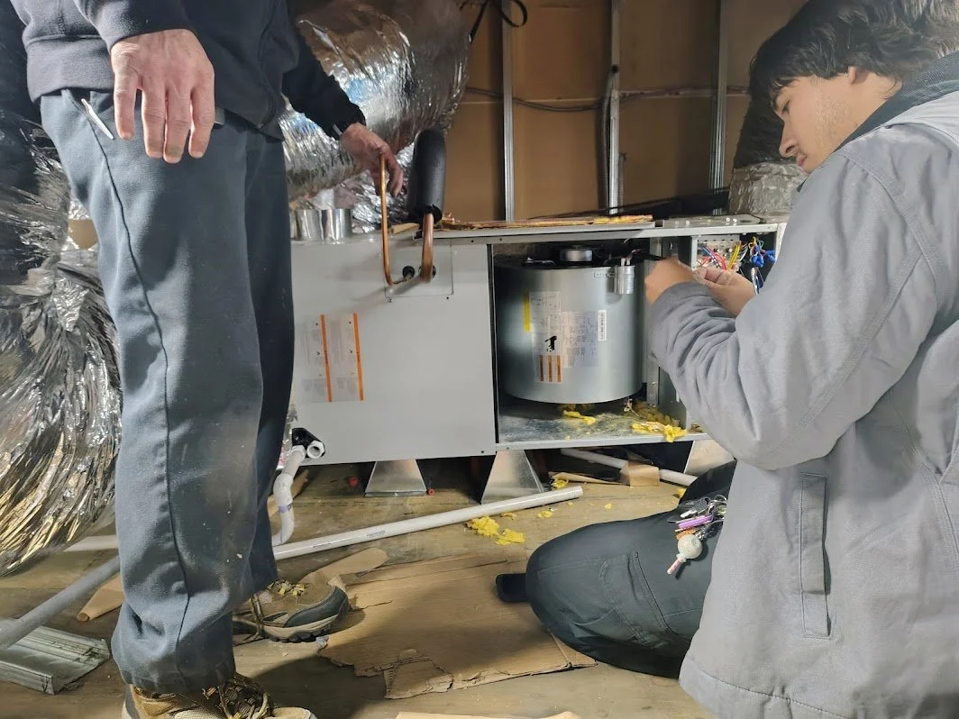 HVACR technicians working on a furnace in a basement with insulation and ductwork, one standing and one kneeling.