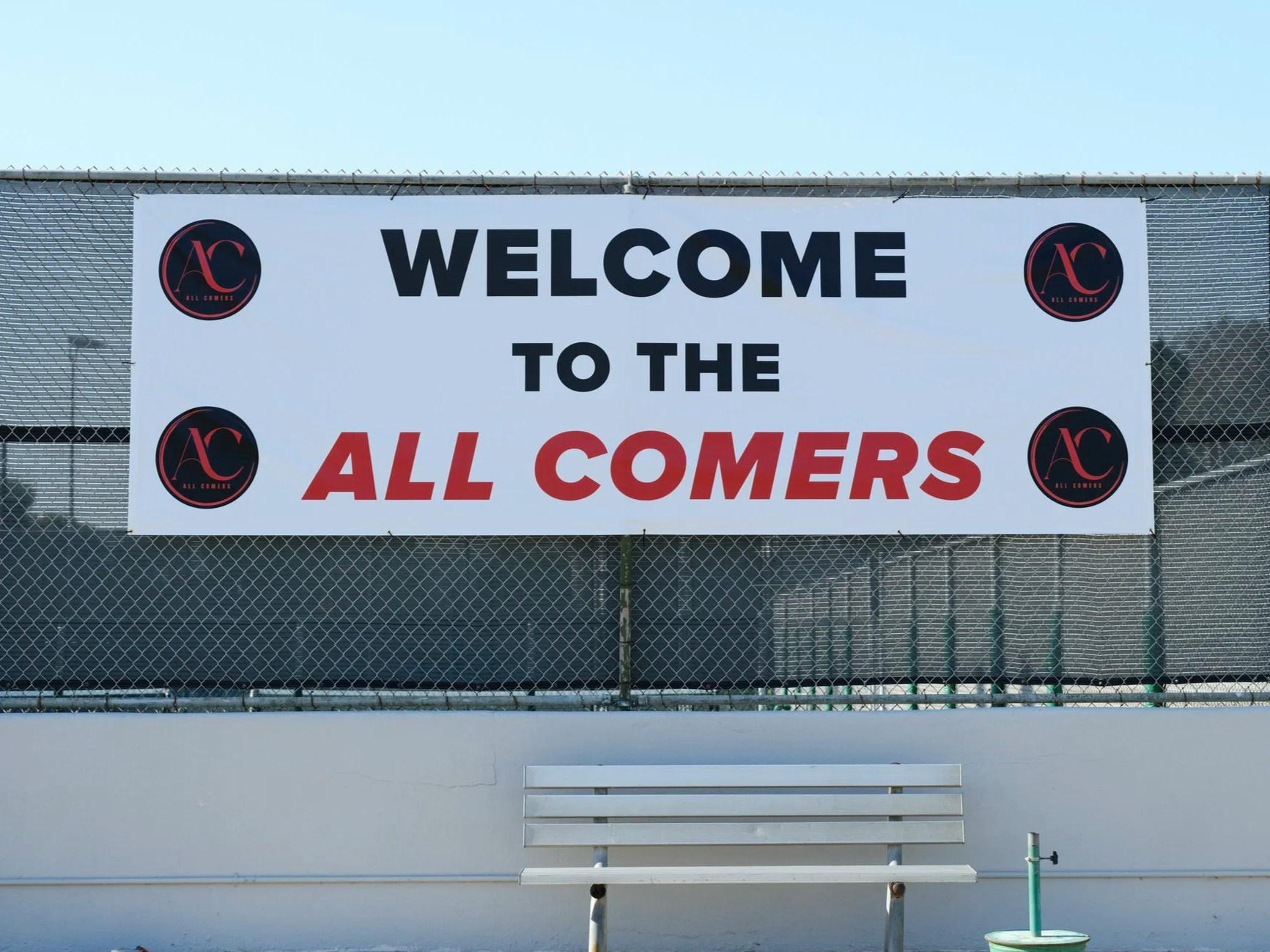 Large welcome sign on a fence reading 'Welcome to the All Comers', with black and red logo for All Comers in each corner, and a white bench in front.