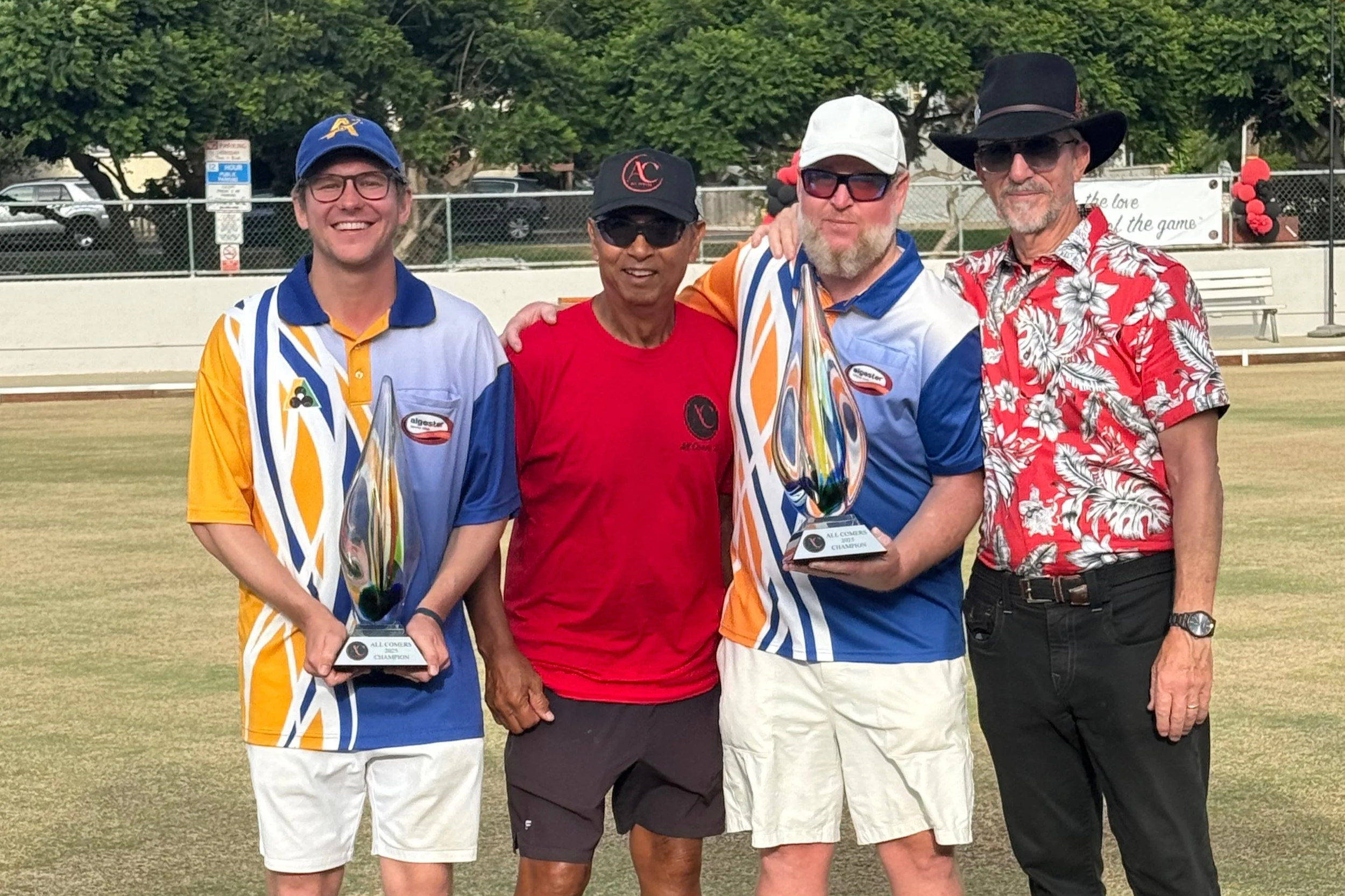 Four men standing on a field, two holding glass trophies, smiling at the camera, with trees and a fence in the background.