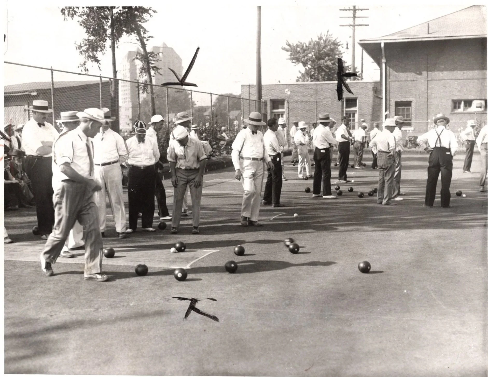 A black and white photo of men playing bocce or bowling on an outdoor court, many dressed in early 20th-century attire with suspenders, hats, and dress shirts, with some sitting on the side and trees and buildings in the background.