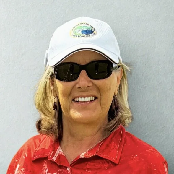 A smiling woman wearing sunglasses, a white baseball cap with a logo, and a red shirt, standing against a plain background.