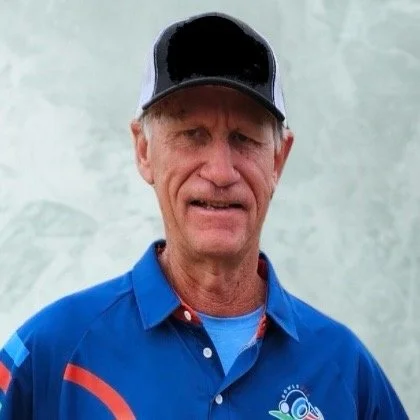 Older man wearing a black and white cap and a blue sports jacket with a logo, standing outdoors.