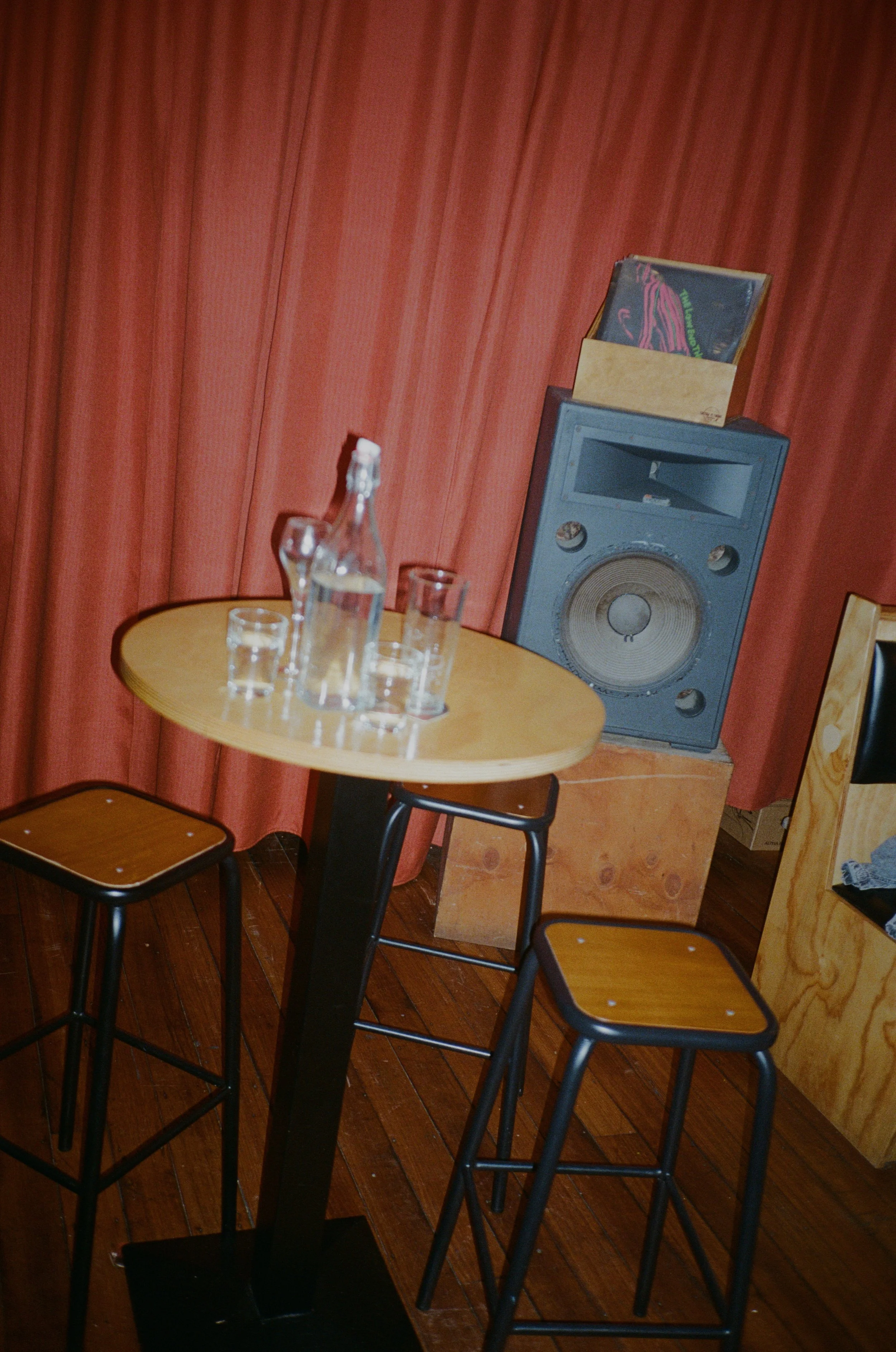 A small round wooden table with several empty glasses and a bottle, two tall stools, a large speaker, and a vinyl record cover, all set against pink curtains.