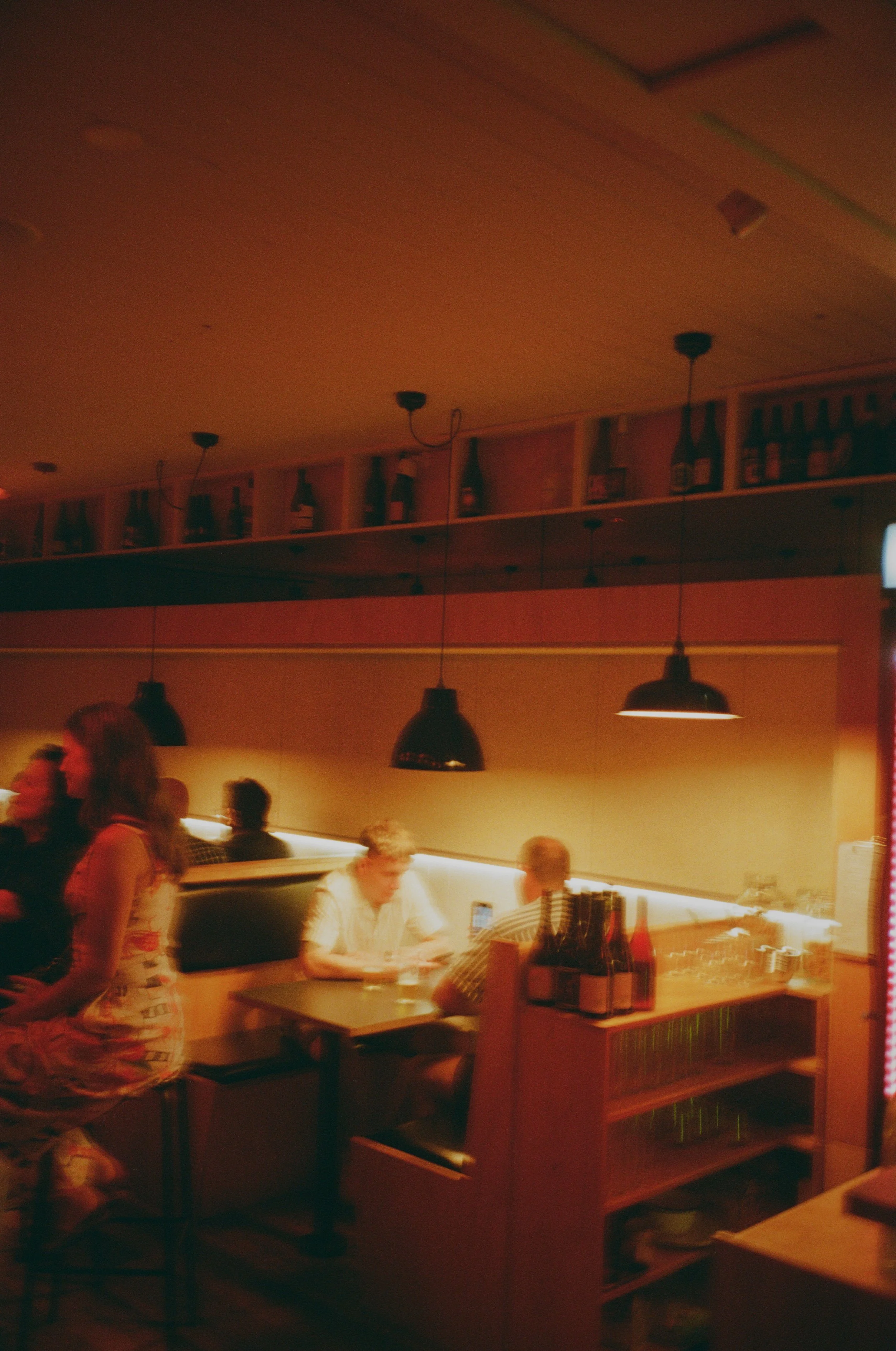Dimly lit restaurant with hanging pendant lights, a bar shelf with bottles on the ceiling, and customers seated at tables, some looking at their phones.